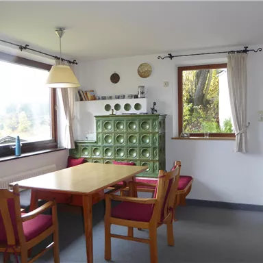 Ein gemütlicher Essbereich mit einem Holztisch und Stühlen in einem hellen Raum. Große Fenster lassen viel Tageslicht herein und bieten einen Blick nach draußen.