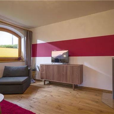 Ein modernes Wohnzimmer mit einem grauen Sofa und einem Holzofen. Ein Fernseher steht auf einem Holzschrank und eine rote Wand sorgt für einen Akzent.
