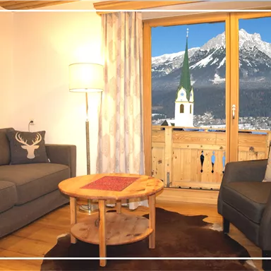 A cozy living room with a gray sofa and a wooden coffee table. Through the window, you can see an impressive mountain landscape and a church tower.