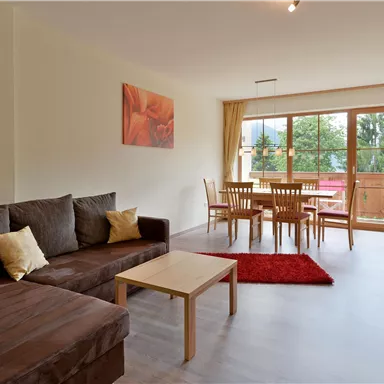 A cozy living room with a couch, a table, and chairs. Large windows provide plenty of daylight and a view outside.