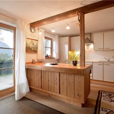 A bright, modern kitchen with wooden details and large windows. The kitchen area features a central cooking island and plenty of space for meal preparation.