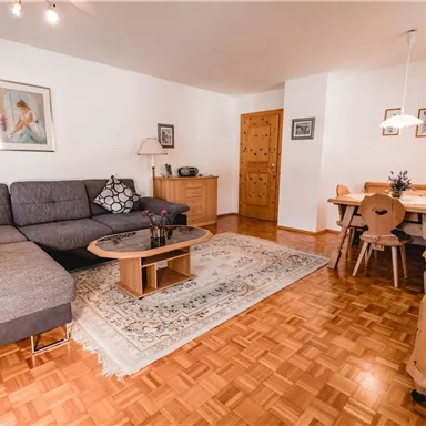 A cozy living room with a large gray sofa and a wooden table. The room is bright and welcoming, with a dining table and decorative elements on the walls.