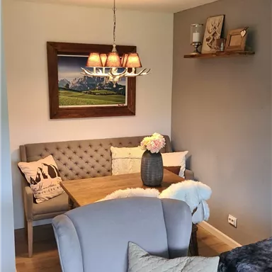 A cozy living area with a bright seating arrangement and a dining table. A landscape painting hangs on the wall, and the lighting creates an inviting atmosphere.