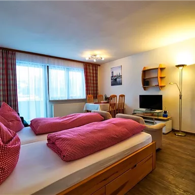 Ein gemütliches Zimmer mit zwei Betten und roten Kissen. Es gibt einen Fernseher, einen Schreibtisch und große Fenster mit natürlichem Licht.