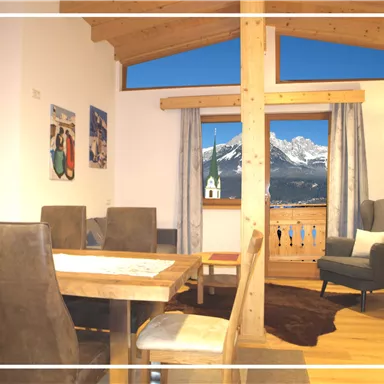 A cozy holiday apartment with wooden decor and a view of the mountains. The space is bright and inviting, with a dining table and a comfortable armchair.
