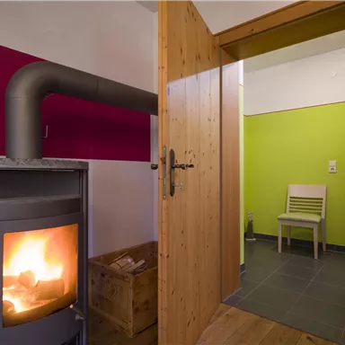 Ein gemütlicher Raum mit einem Holzofen und bunten Wänden. Im Hintergrund ist ein Stuhl auf einem Fliesenboden sichtbar.