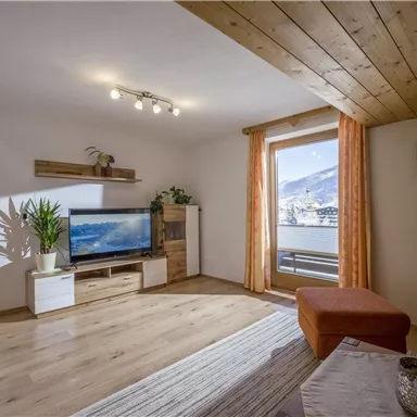 Ein gemütliches Wohnzimmer mit Holzdecke und großen Fenstern. Es gibt eine moderne Fernsehecke und Pflanzen, die eine einladende Atmosphäre schaffen.