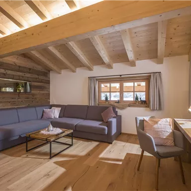 Ein moderner Wohnraum mit einem bequemen grauen Sofa und einem Holzstuhl. Große Fenster sorgen für viel Tageslicht und eine warme Atmosphäre.