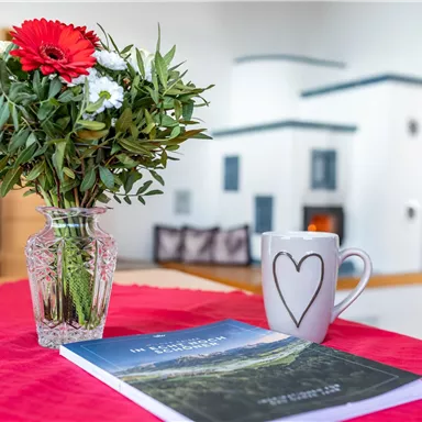 Ein gedeckter Tisch mit einer Vase voller Blumen und einer Tasse Kaffee. Im Hintergrund liegt ein Buch mit einer Naturlandschaft auf einem roten Tischläufer.
