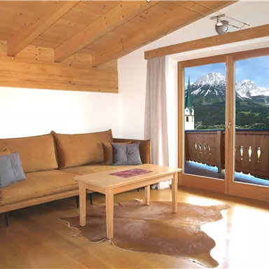 A cozy living room with a sofa and a wooden table. Large windows offer a beautiful view of the mountains.