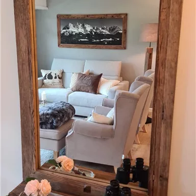 A large wooden mirror with a shelf. In the background, a cozy living room is visible with a bright sofa and wall decoration.
