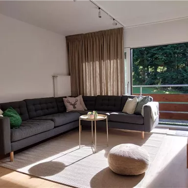 A cozy living room with a gray sofa and a small table. Large windows offer a view of the green trees outside.