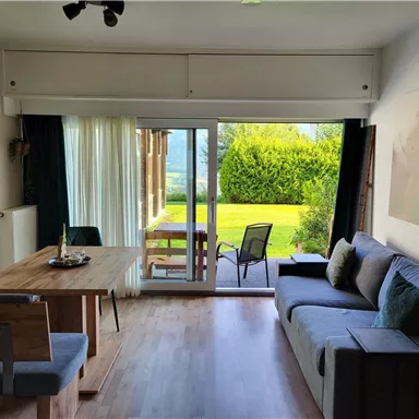 Ein gemütlicher Wohnraum mit einem Essbereich und einem Sofa. Große Fenster bieten einen Blick auf den Garten im Freien.