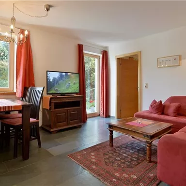 A cozy living room with a red upholstered sofa and a wooden table. Large windows let in plenty of light and offer a view of the nature outside.