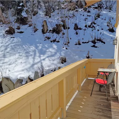 A wooden balcony with a table and two chairs overlooks a snow-covered landscape. The sun shines on the snowy area in the background.