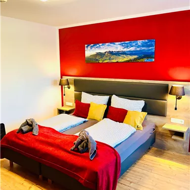 A modern bedroom with a large double bed and colorful pillows. A landscape painting hangs on the wall, and the room has a cozy ambiance.