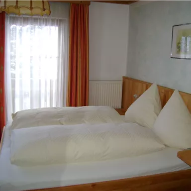 A cozy bedroom with a double bed and natural wood furniture. Bright curtains and orange window decorations create a warm atmosphere.