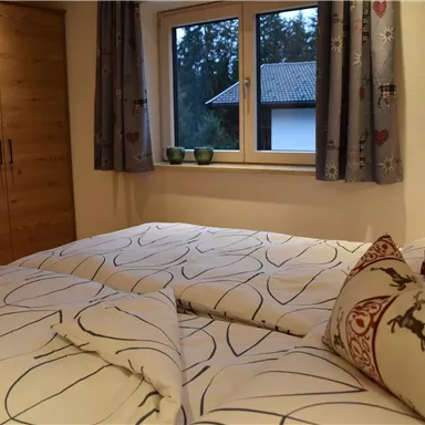 A cozy bedroom with a large window and a wooden wardrobe. The bed is adorned with white patterned blankets and decorative cushions.