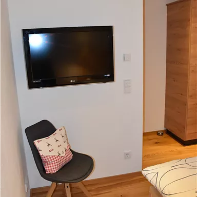 A cozy room with a TV on the wall and a chair with a cushion. The floor is wooden and the furniture is modern.