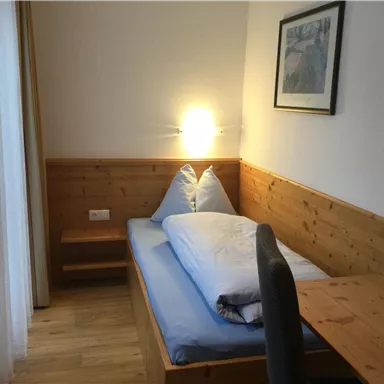 A cozy room with a single bed, a desk, and beautiful wooden details. The walls are bright, and there is a window with curtains.