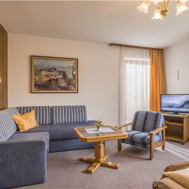Ein gemütliches Wohnzimmer mit einer blauen Couch und einem kleinen Tisch. Große Fenster mit Vorhängen bringen viel Licht in den Raum.