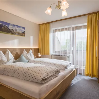 Ein gemütliches Schlafzimmer mit zwei Betten und großen Kissen. Helle Fenster und gelbe Vorhänge schaffen eine freundliche Atmosphäre.