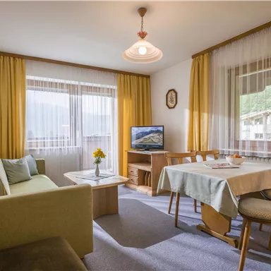 Ein gemütliches Wohnzimmer mit einem Sofa, einem Tisch und Stühlen. Große Fenster lassen viel Licht herein und bieten einen Blick auf die Umgebung.