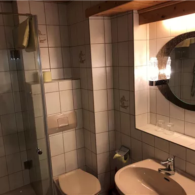 A simple bathroom with shower, sink, and mirror. The walls are tiled and the furnishings are functional.