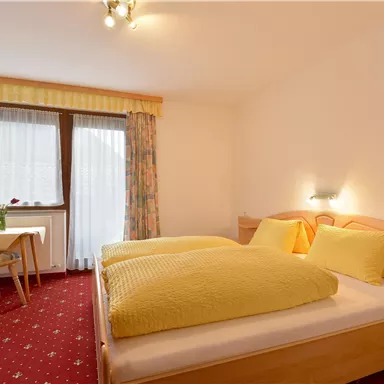 Ein gemütliches Schlafzimmer mit zwei Betten und gelben Kissen. In der Ecke steht ein Tisch mit Stühlen und großen Fenstern, die für viel Licht sorgen.