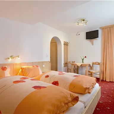 Ein gemütliches Schlafzimmer mit zwei Betten und dekorativen Kissen. Der Raum hat einen Fernseher und einen kleinen Esstisch sowie helle Vorhänge.