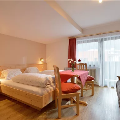 Ein einfaches Zimmer mit zwei Betten, einem Esstisch und einer Schlafcouch. Große Fenster sorgen für viel Tageslicht und eine angenehme Atmosphäre.
