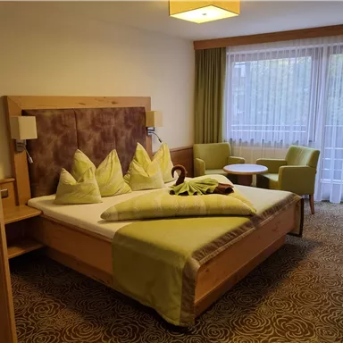 Ein modernes Hotelzimmer mit einem Doppelbett, frischen Farben und gemütlichen Stühlen. Großer Fensterbereich mit viel Tageslicht.