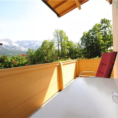 A balcony with a table and a chair. In the background, trees and mountains can be seen.