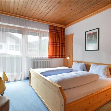 A cozy room with a double bed and wooden decor. Large windows provide plenty of daylight and a pleasant atmosphere.