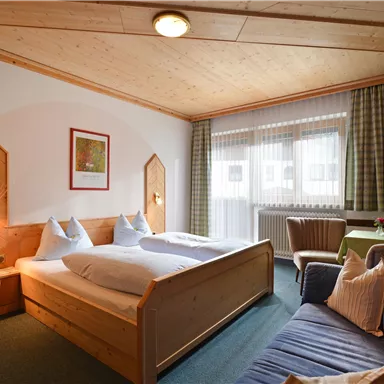 A cozy room with two beds and wooden decor. There is a comfortable armchair and plenty of daylight through the windows.