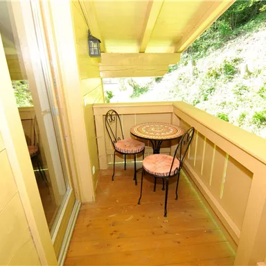 A small balcony with a table and two chairs. The surroundings consist of lush greenery.