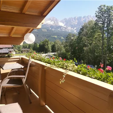 A beautiful balcony with a view of the mountains and green trees. Flowers adorn the railing and the sun is shining.