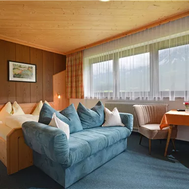 A cozy room with wooden paneling and large windows. There is a comfortable sofa, cushions, and a small table with chairs.