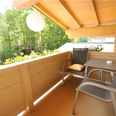 A cozy balcony with a table and two chairs. The view overlooks a green, wooded area.
