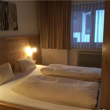 A cozy bedroom with a double bed and two pillows. The warmly lit room features wooden details and large windows with curtains.