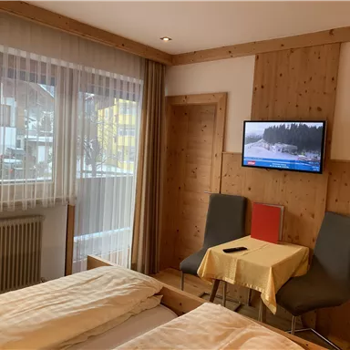 A cozy hotel room with two beds and a TV on the wall. There are two chairs at a small table and plenty of daylight coming in through the windows.