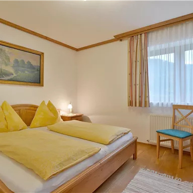 A bright bedroom with a double bed and yellow pillows. There is a window with curtains and a chair next to the bed.