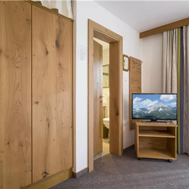 Ein gemütliches Zimmer mit einem Holzwandschrank und einem Fernseher. Durch die Tür auf der rechten Seite ist das Badezimmer sichtbar.