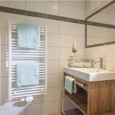 Ein modernes Badezimmer mit einem weißen Waschbecken und einem Holzschrank. An der Wand hängen zwei grüne Handtücher und ein Handtuchwärmer ist vorhanden.