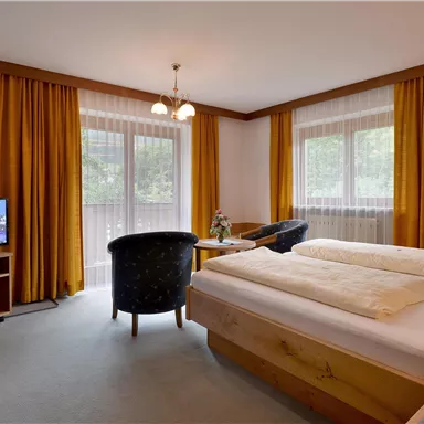 Ein gemütliches Hotelzimmer mit zwei Betten und einer Sitzecke. Große Fenster lassen viel Licht herein, und ein Fernseher ist ebenfalls vorhanden.