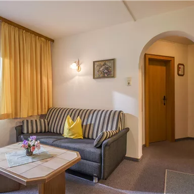 Ein gemütliches Wohnzimmer mit einem grauen Sofa und einem kleinen Tisch. Der Raum hat große Fenster mit Vorhängen und eine Tür, die zu einer anderen Wohnung führt.