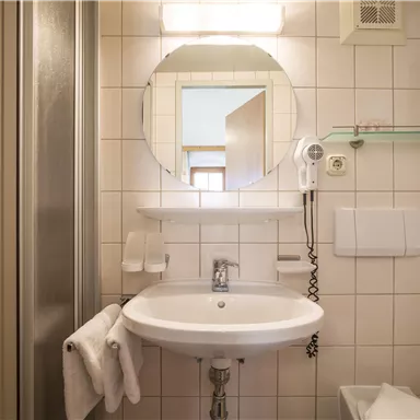 A modern bathroom with a shower, a sink, and a large mirror. The walls are tiled and there are some towels and a hairdryer.