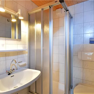 Ein modernes Badezimmer mit einer Dusche, einem Waschbecken und einem großen Spiegel. Die Wände sind gefliest und die Beleuchtung ist hell.