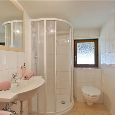 Ein modernes Badezimmer mit einer Duschkabine, einem Waschbecken und einer Toilette. Die Wände sind hell gefliest, und es gibt Handtücher in sanften Farben.