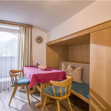 Ein gemütlicher Essbereich in einer hellen Küche. Der Tisch ist mit einer roten Tischdecke gedeckt und es gibt Stühle aus hellem Holz.